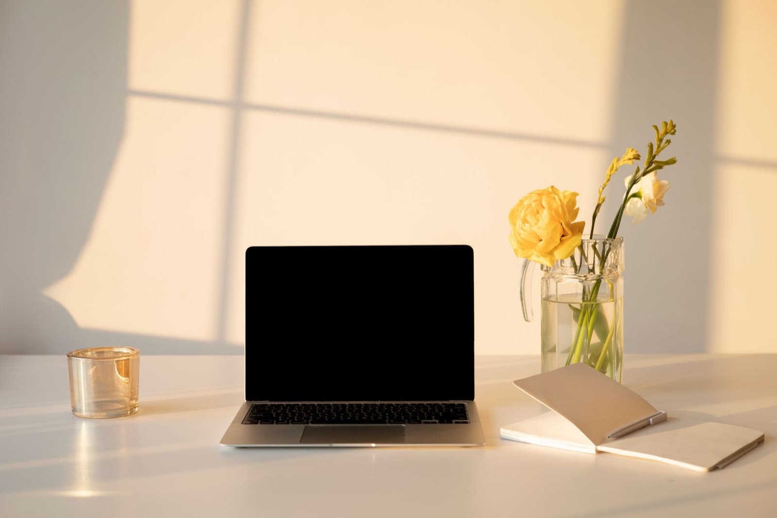 Laptop on a white desk in warm morning light beside a glass vase of yellow flowers, a candle holder and an open notebook