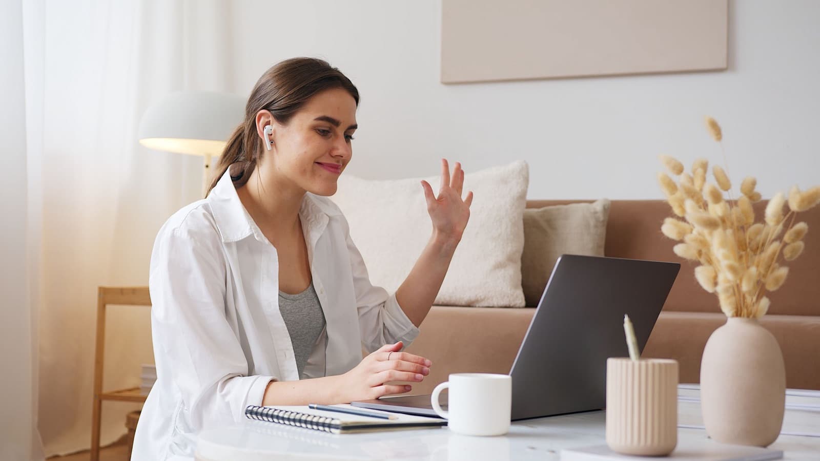 Smiling young woman with earbuds waving at her laptop during a video call in a bright, neutral living room