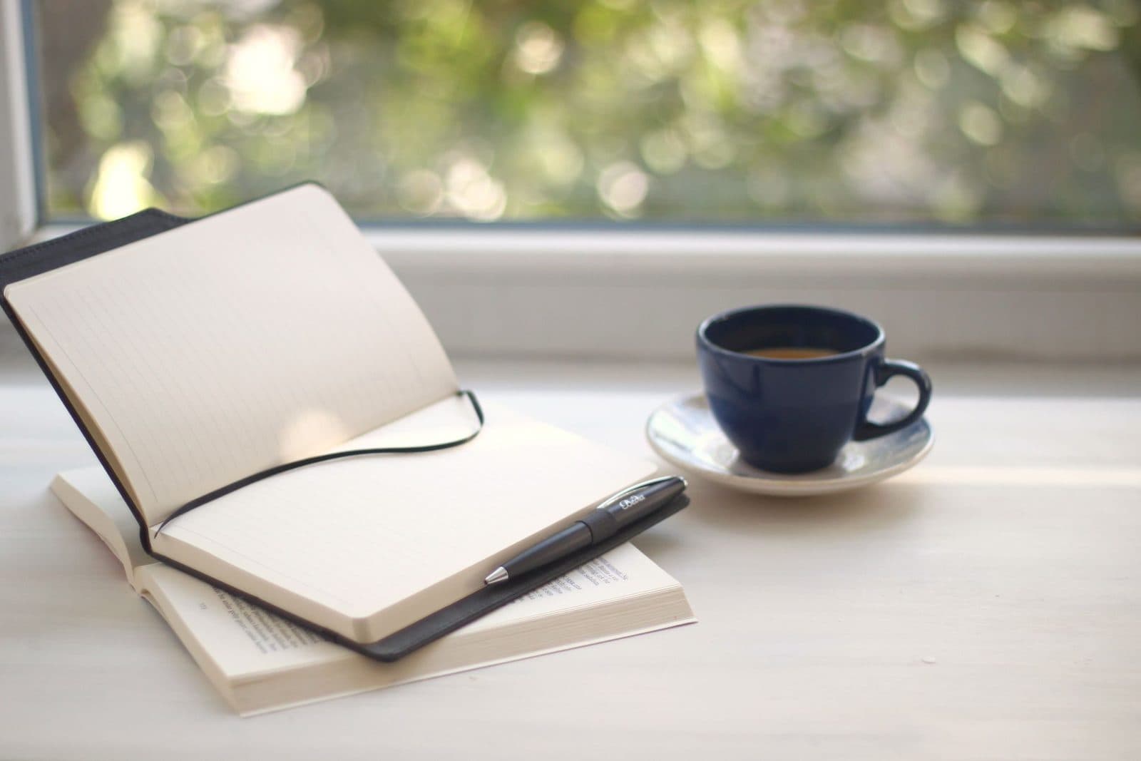 Open notebook and pen beside a cup of coffee on a bright windowsill