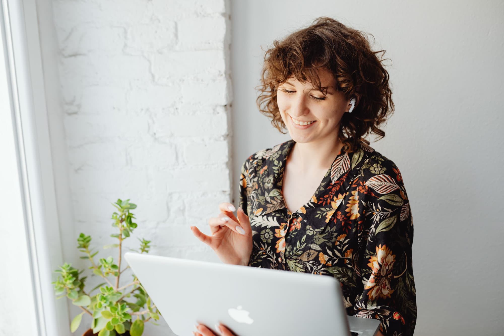 Smiling woman with curly hair and wireless earbuds using a laptop beside a window and a potted plant