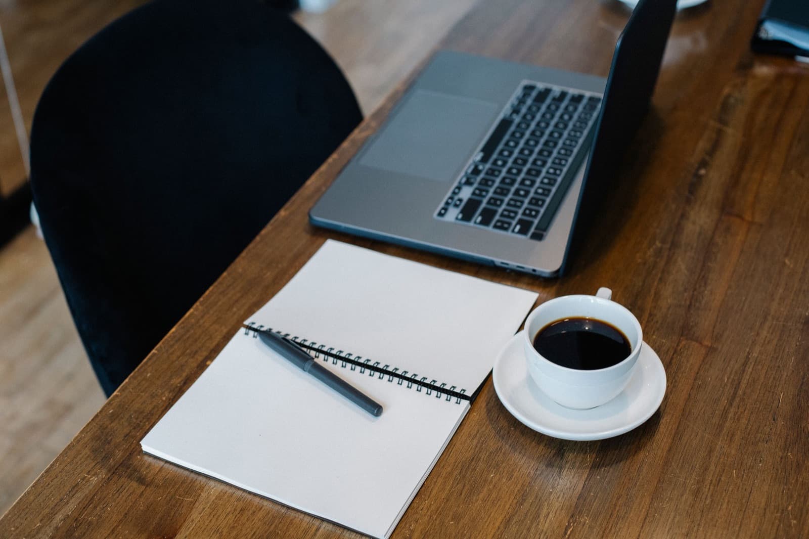Open blank spiral notebook with a pen beside a cup of black coffee and a laptop on a wooden table