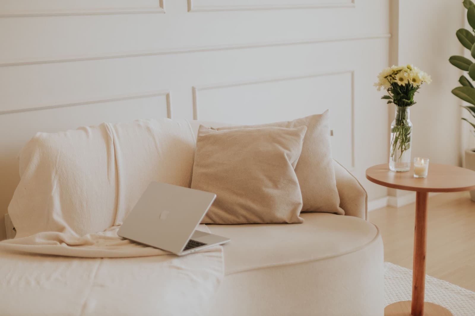 Cream sofa with pillows and an open laptop beside a small wooden side table holding a vase of yellow daisies and a candle