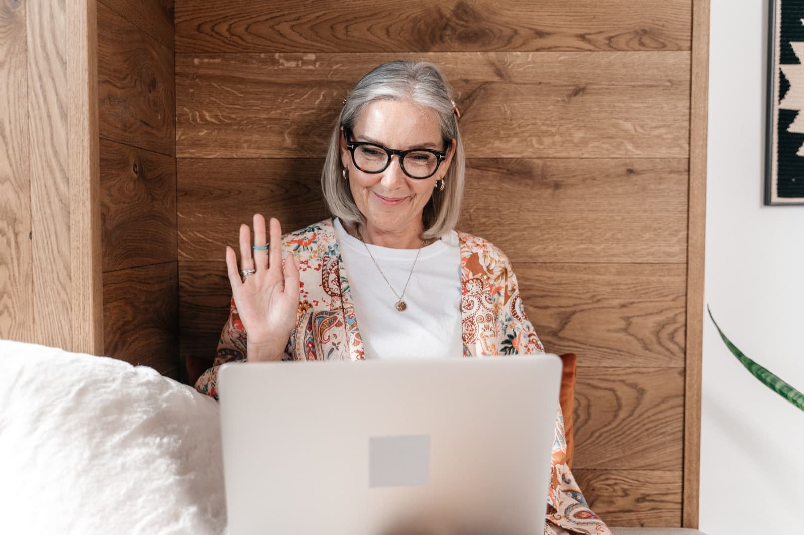 Smiling older woman with gray hair and glasses waving at her laptop during a video call at home