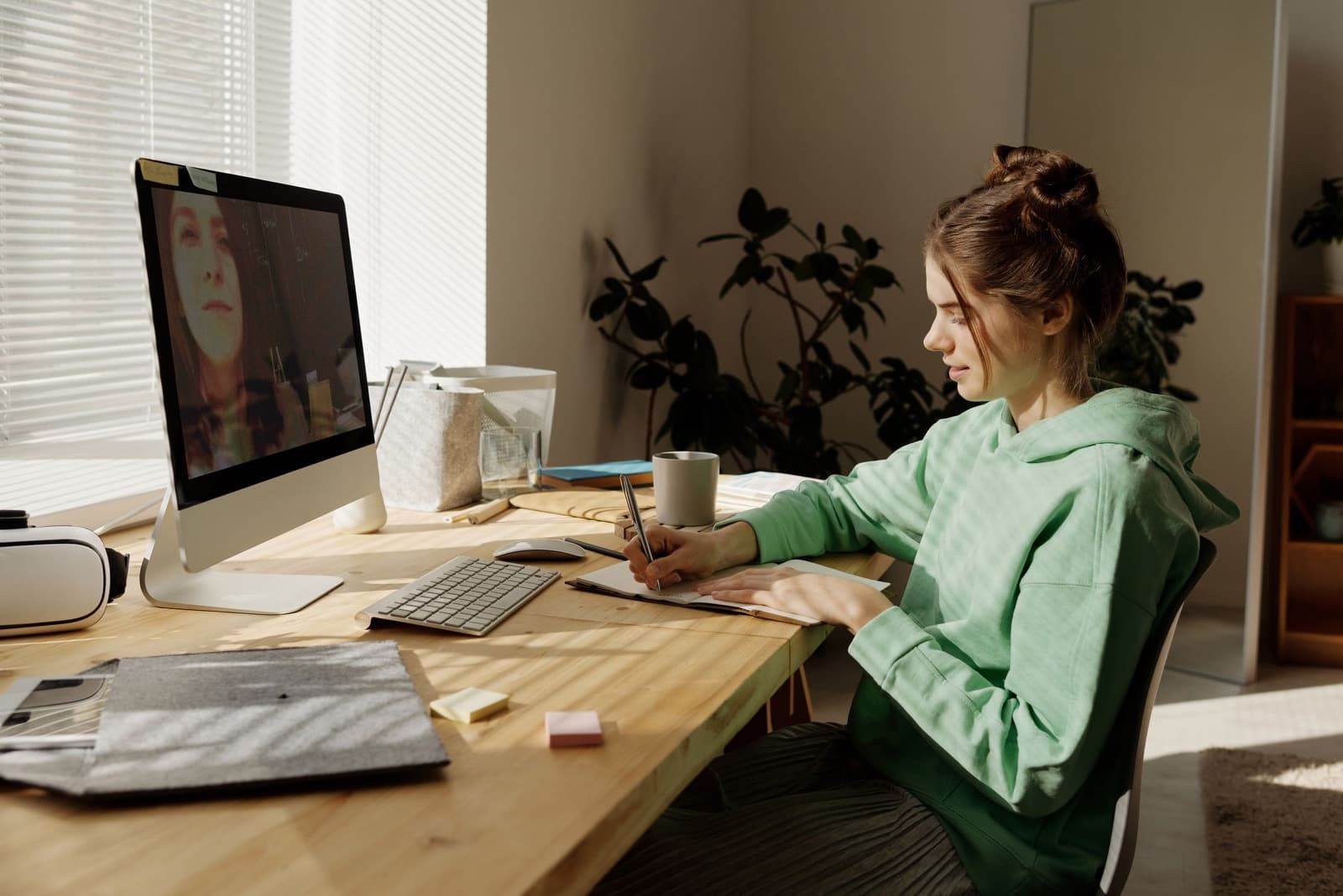 Teenage girl in a mint green hoodie taking notes at a sunlit desk while on a video call on a desktop computer